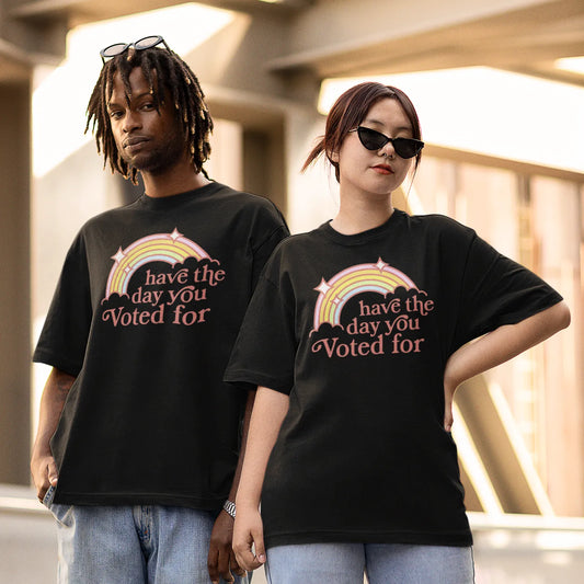 Two people wearing unisex black t-shirts with a colorful graphic with a rainbow and sparkles that says, “ Have The Day You Voted For’ Rebel Girl Rampage, Anne Lesniak