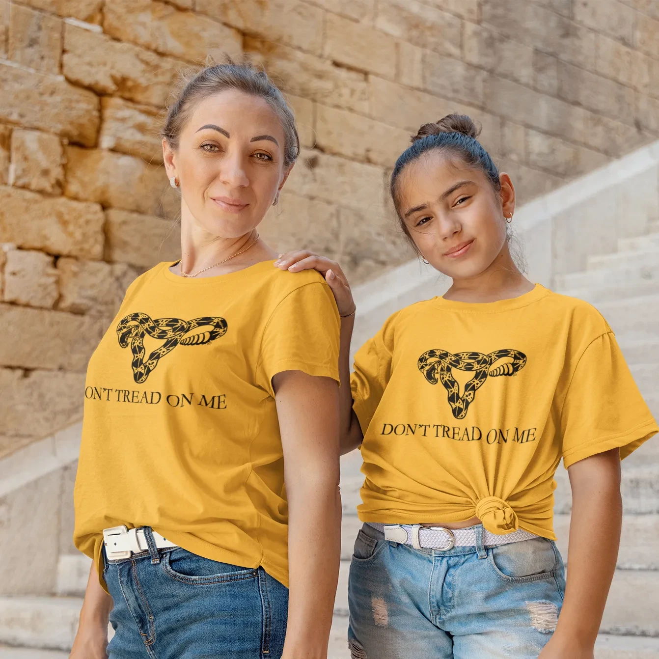 Two women wearing gold triblend Don’t Tread On Me Uterus T-shirts outside on a staircase, Women’s Rights, Anne Lesniak, Rebel Girl Rampage 