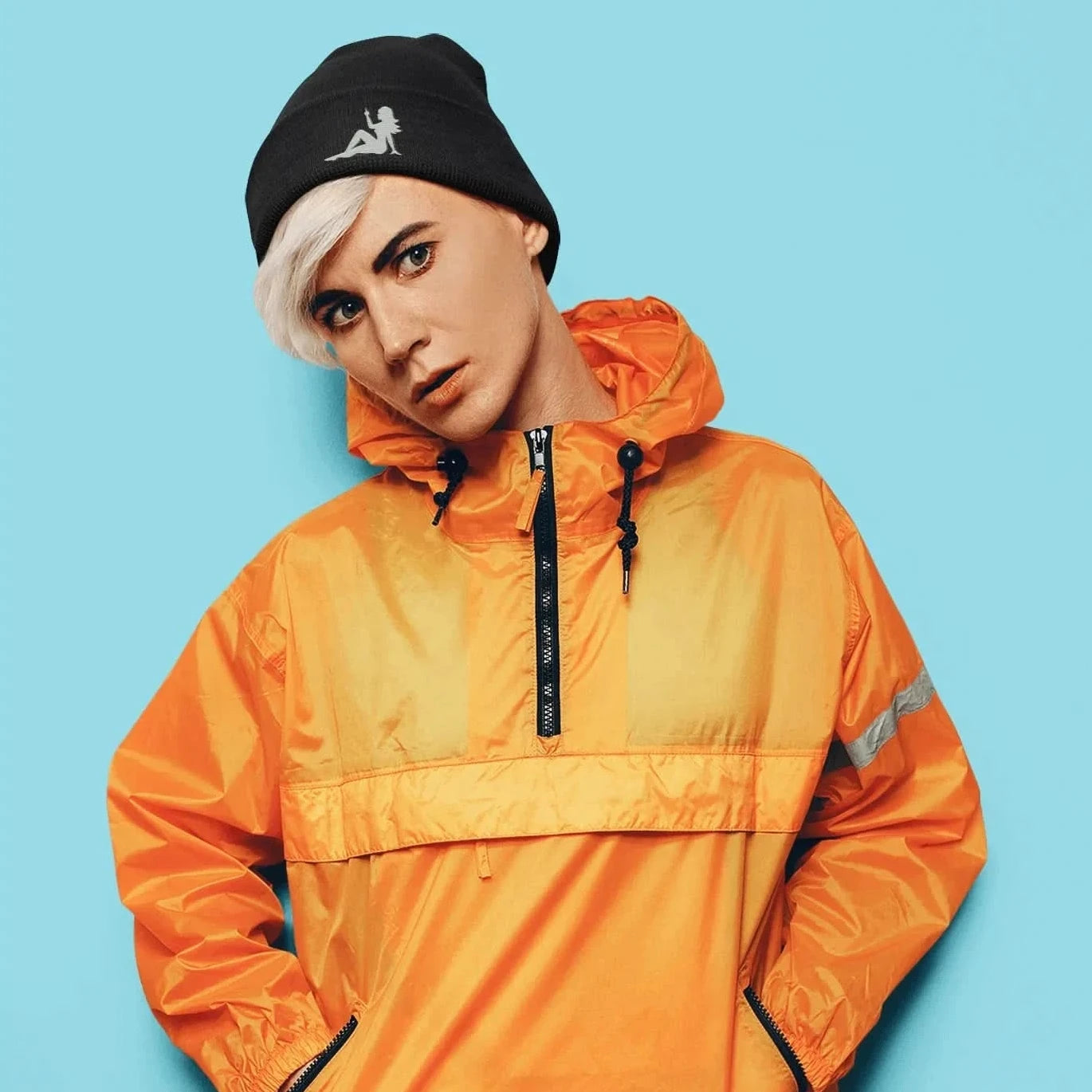 Person wearing an orange jacket and black mudflap girl flipping the bird embroidered beanie against a blue background