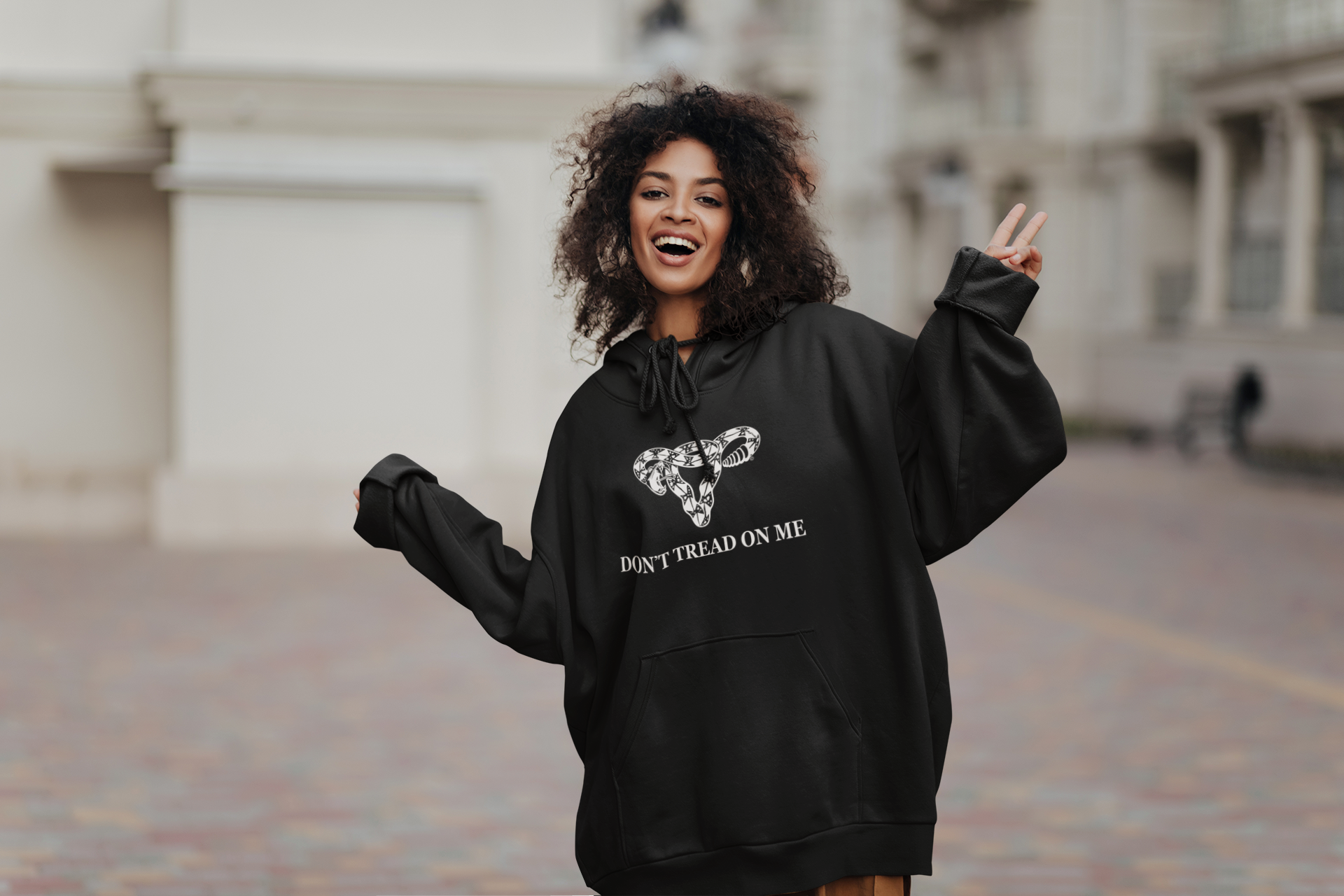 Happy woman wearing an oversized black unisex hoodie with a Don’t tread on e m uterus rattlesnake design giving the peace sign standing outdoors. Rebel Girl  Rampage